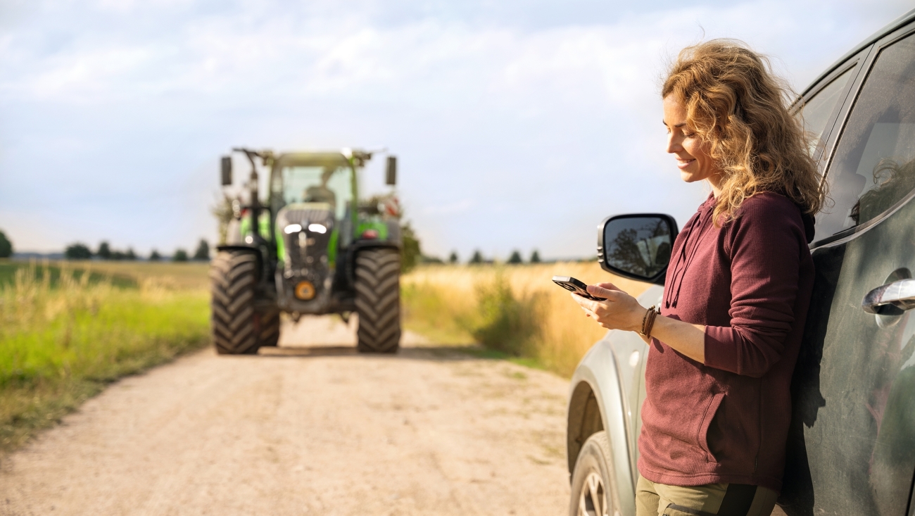 Eine Landwirtin lehnt sich an ihrem Auto an und liest den Fendt Newsletter auf dem Handy.