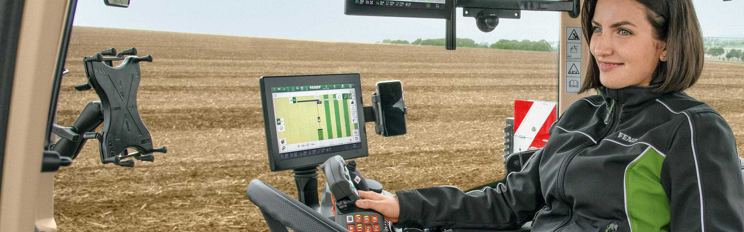 Ein Landwirtin sitzt in der Fendt 700 Gen 7 Kabine. Im Hintergrund in der Innenaustattung ist ein Tablet auf dem FendtOne Onboard geöffnet ist.