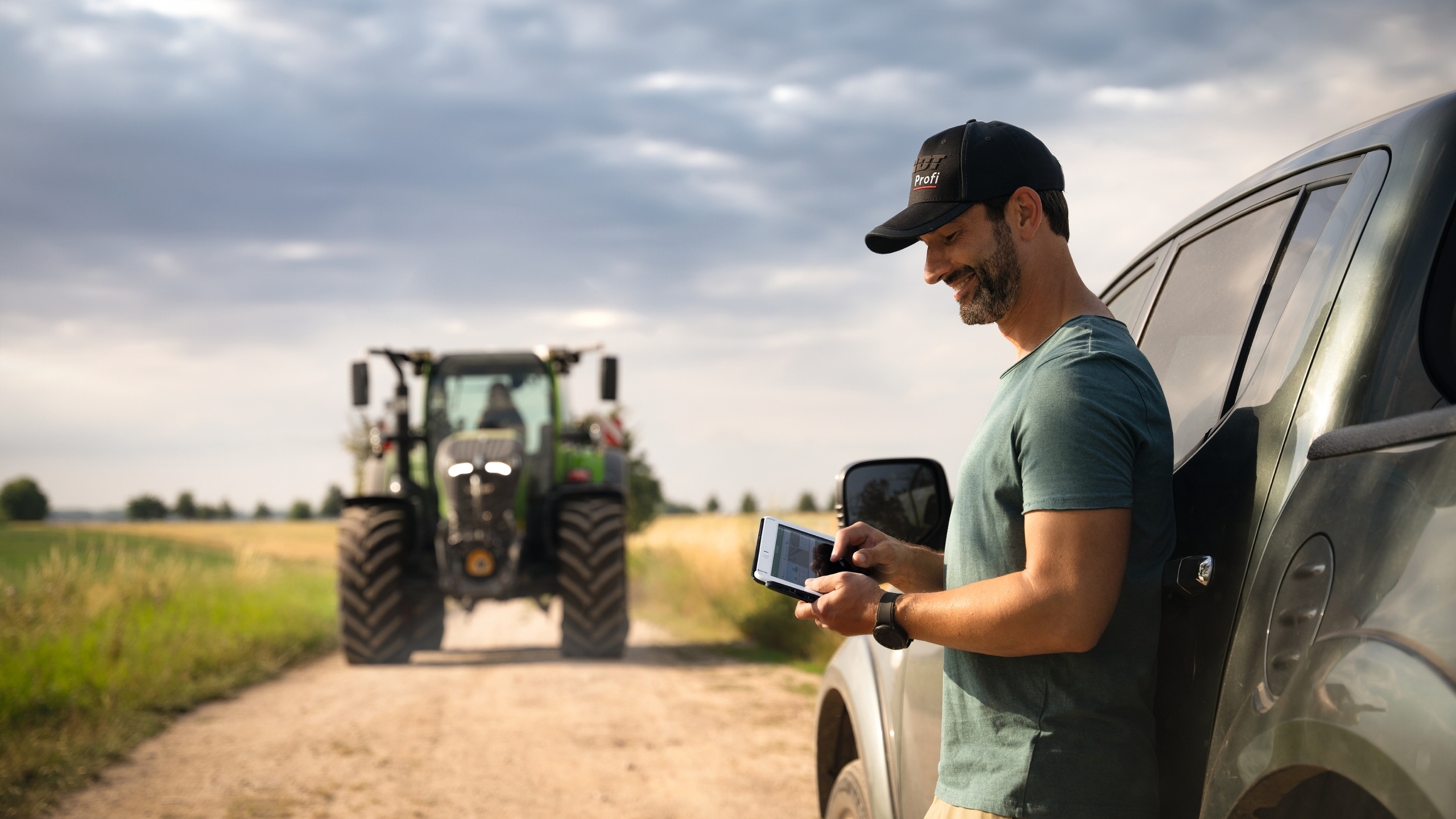 Ein Landwirt hält ein Tablet in der Hand und lehnt sich an sein Auto an. Im Hintergrund kommt ein Fendt 700 Vario Gen7 angefahren.