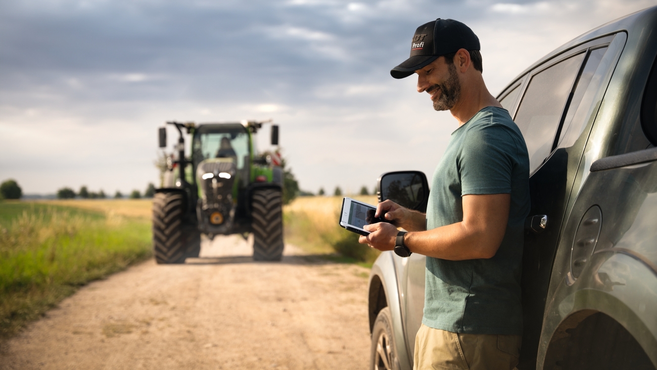 FendtONE offboard Ein Landwirt hält ein Tablet in der Hand und lehnt sich an sein Auto an. Im Hintergrund kommt ein Fendt 700 Vario Gen7 angefahren.