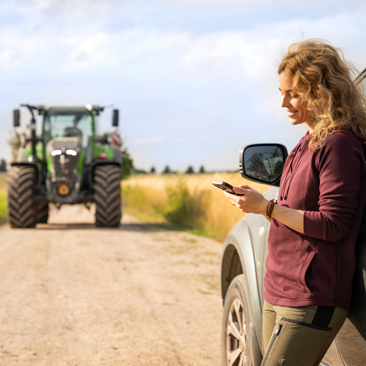 Eine Landwirtin hält ein Tablet in der Hand und lehnt sich an sein Auto an. Im Hintergrund kommt ein Fendt 700 Vario Gen7 angefahren.
