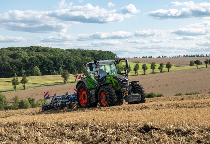 Ein Fendt Traktor mit einem Grubber auf einem Feld mit blauem Himmel und Wolken im Hintergrund.