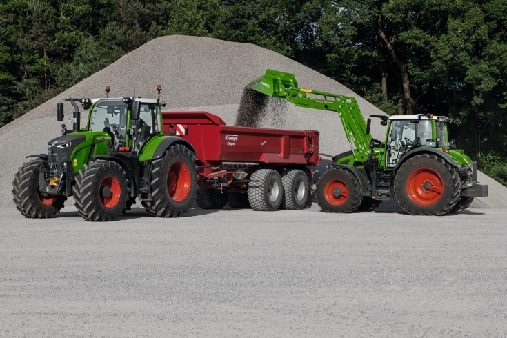 Ein Fendt Traktor lädt Kies auf einen roten Anhänger. Der Anhänger hängt an einem zweiten Fendt Traktor