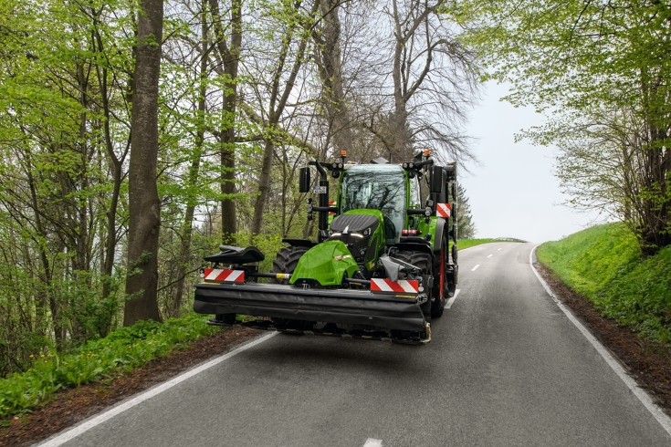 Ein Fendt Traktor mit einem Fendt Mähwerk fährt auf einer Landstraße durch den Wald.