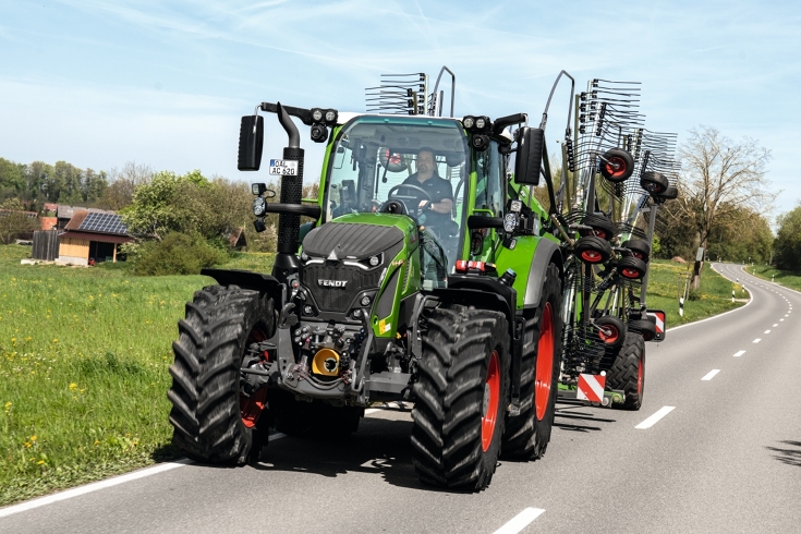 Ein Fendt Traktor mit einem angehängten Fendt Schwader fährt auf einer Landstraße.