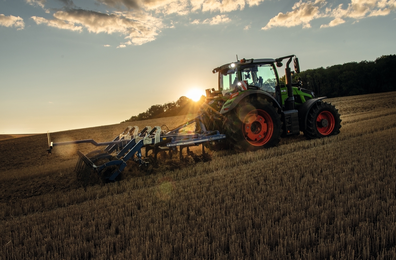 Ein Fendt Traktor mit einem Grubber fährt bei Sonnenuntergang über ein Stoppelfeld