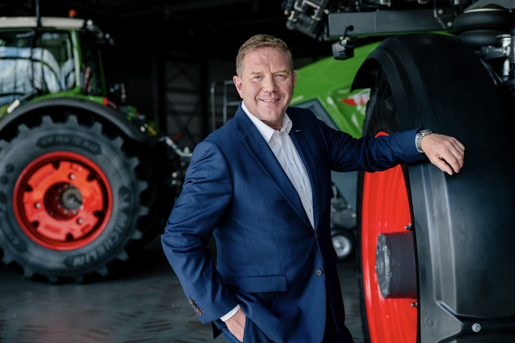 Ein Mann steht in einer Halle und lehnt an einem Fendt Traktor. Im Hintergrund sieht man einen weiteren Traktor mit einer Ballenpresse.