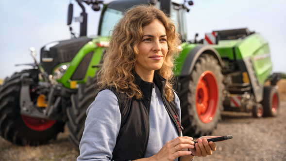 Eine Landwirtin vor ihren Fendt Landmaschinen öffnet die Fendt News App auf ihrem Handy