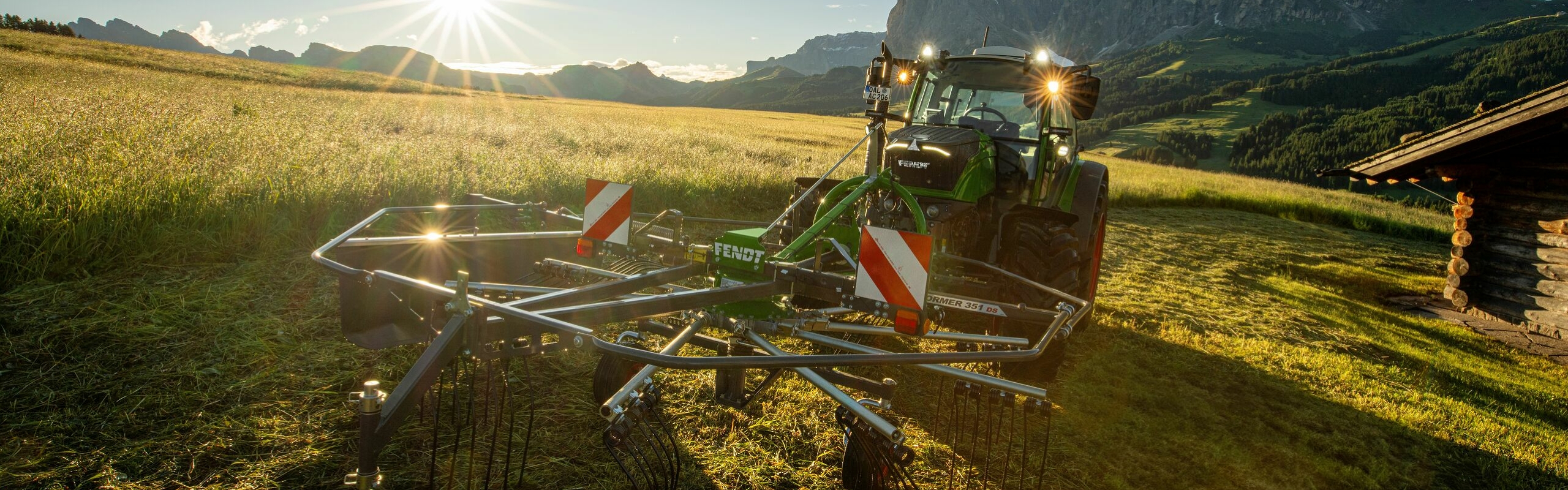 Der grüne Fendt Traktor mit roten Felgen und dem grünen Anbaugerät Fendt Former Alpin im Einsatz auf einer Bergwiese. Im Hintergrund ist eine Berglandschaft, ein blauer Himmel und die strahlende Sonne.