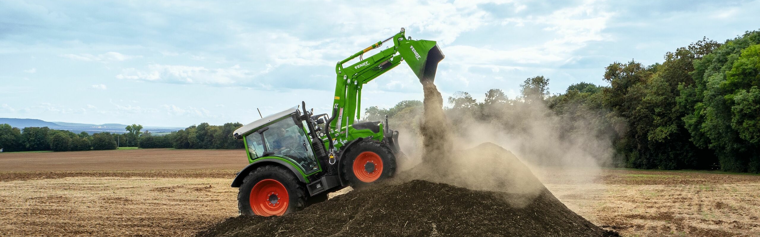 Fendt 200 Vario  auf dem Acker mit Fendt Cargo und schüttet Erde auf. Im Vordergrund sind Staub und im Hintergrund Bäume zu sehen.