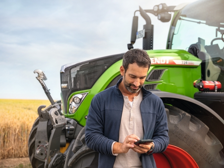 Ein Landwirt steht neben einem Fendt 724 Traktor und blickt auf sein Handy.