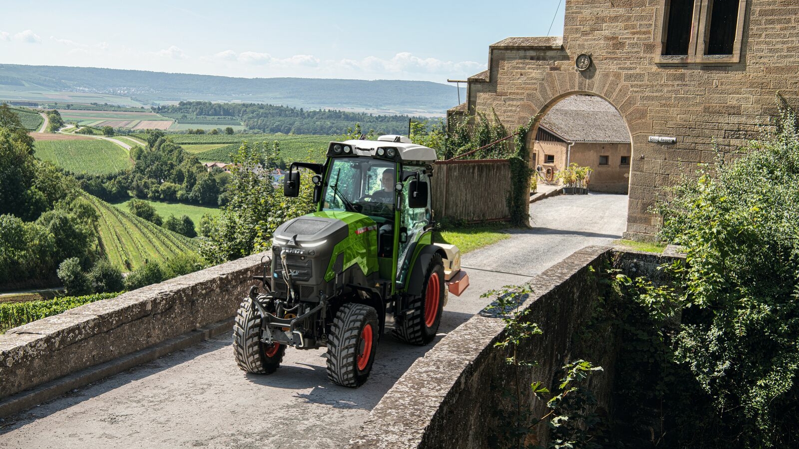 Der Fendt e100V Vario Ein grüner Fendt e100 V Vario fährt aus einem Hof oberhalb der Weinberge über eine Brücke