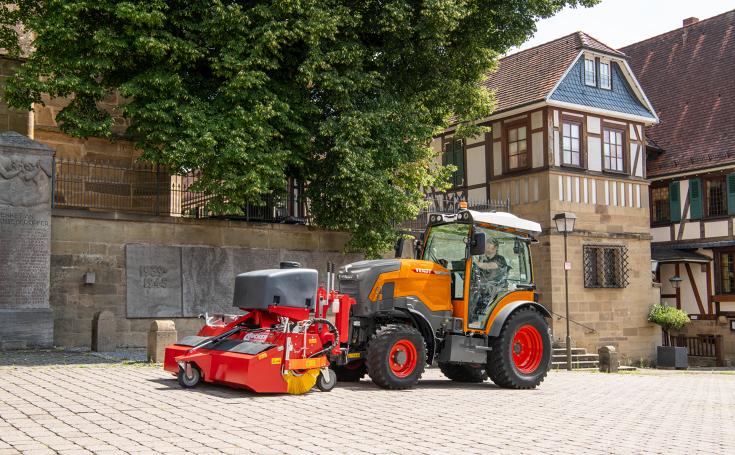 Fendt e107 V Vario im Kommunaleinsatz mit Kehrmaschine Ein kommunal-orange gefärbter Fendt e107 V Vario mit einer Kehrmaschine an der Front kehrt das Pflaster in einer Stadt, im Hintergrund Mauern mit Sandstein, ein Baum und ein Fachwerkhaus