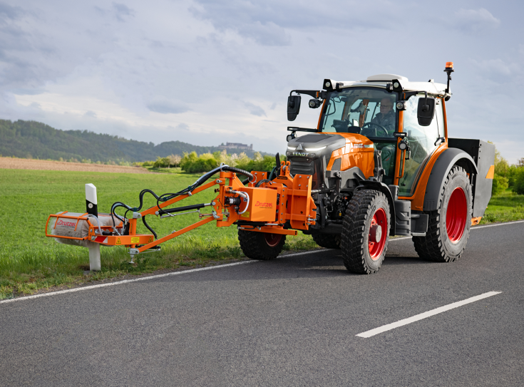 Ein Fendt e107 Vario bei der Reinigung von Leitpfosten Ein Fendt e107 Vario in kommunal-orange mit einem Frontanbaugerät zur Reinigung von Leitpfosten fährt auf der Straße und reinigt Leitpfosten
