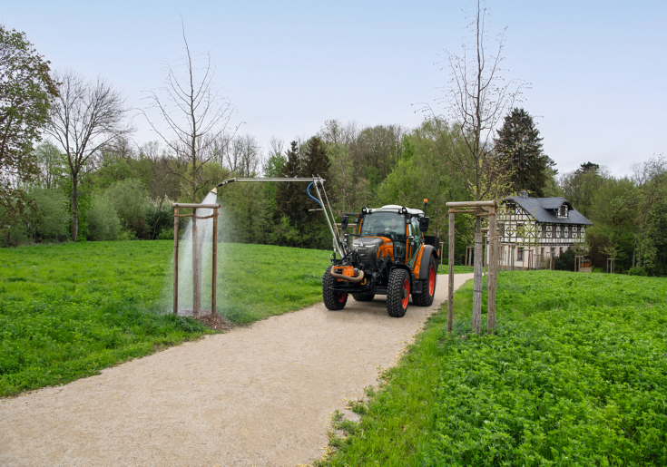 Fendt e100 Vario beim Bewässern Ein Fendt e107 Vario in kommunal-orange bewässert mit einem an der Front des Traktors angebauten Gießarm einen Baum in einem Park im Frühjahr, im Hintergrund ist ein altes, renoviertes Fachwerkhaus zu sehen