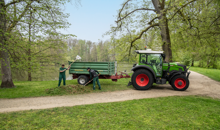 Fendt e100 Vario mit Anhänger im Park Zwei Mitarbeitende in der Landschaftspflege beladen einen Anhänger hinter einem Fendt e107 Vario mit Blättern in einem Park mit einem von Bäumen umringten See