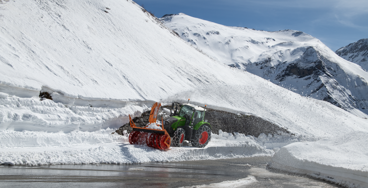 Fendt 728 Vario mit Schneeketten im Winterdienst Ein Fendt 728 Vario in grün fährt mit Schneeketten und einer Schneefräse durch hohen Schnee um die Straße freizufräsen im Hintergrund verschneite Berge