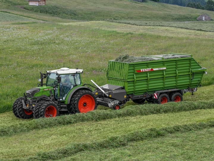 Ein grüner Fendt Traktor mit roten Felgen und einem grünen Fendt Tigo Ladewagen mit roten Felgen fährt auf einer grünen Bergwiese