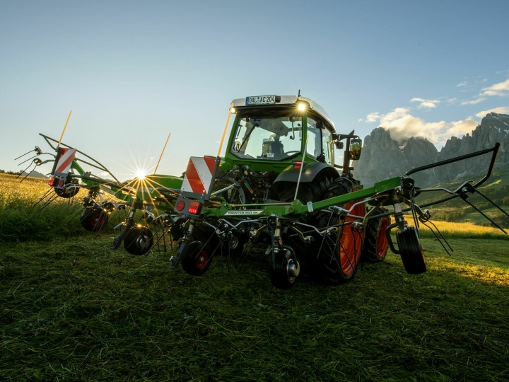 Auf einer Bergwiese steht der grüne Heuwender Fendt Twister vor einem grünen Fendt Traktor. Im Hintergrund geht die Sonne langsam unter, der Himmel ist blau und die Berge sind ebenfalls zu sehen