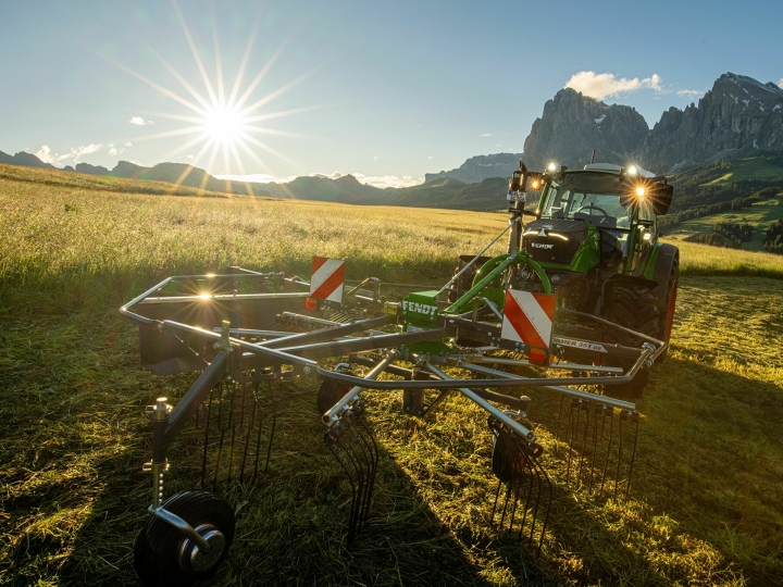 Auf einer Bergwiese steht der grüne Schwader Fendt Former Alpin vor einem grünen Fendt Traktor. Im Hintergrund geht die Sonne langsam unter, der Himmel ist blau und die Berge sind ebenfalls zu sehen