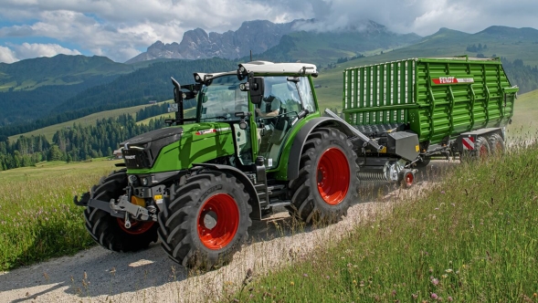 Ein grün lackierter Fendt 200 Vario mit angehängtem Fendt Tigo Ladewagen fährt im alpinen Raum über einen Feldweg während der Grünfutterernte.