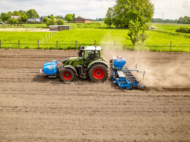 Ein grüner Trakor mit roten Felgen fährt mit einer blauen Sähmaschine über den Acker