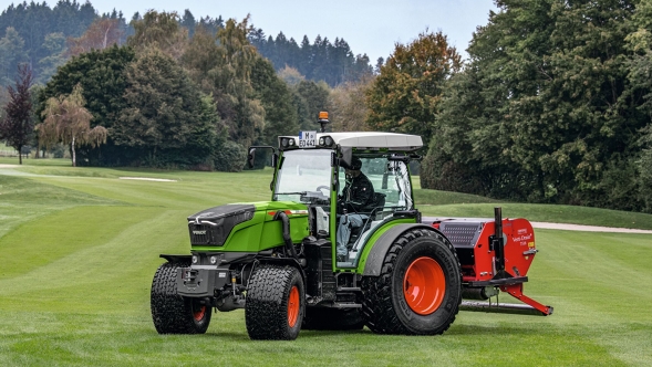 Der grüne Traktor mit den roten Felgen fährt mit einem roten Anbaugerät über einen Golfplatz.