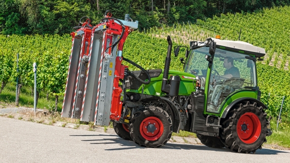 Der grüne Fendt 200 V/F/P mit den roten Felgen fährt mit einer roten Schneidemaschine für den Weinbau auf einer Straße durch die Weinberge.