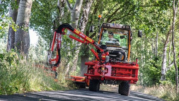 Der orangene Fendt fährt mit einem roten Mulcher auf der Straße und mulcht den Randstreifen.