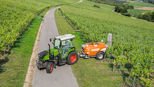 Ein grüner Fendt 200 V/F/P Vario mit roten Felgen fährt mit einer orangenen Pflanzenschutzspritze aus dem Weinberg auf eine Straße.