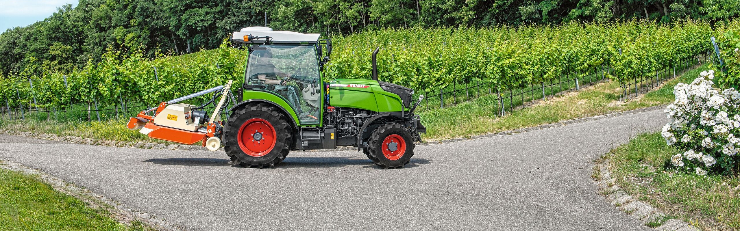 Der grüne Fendt 200 V/F/P Vario mit den roten Felgen fährt auf der Straße. Er hat einen orangenen Mulcher als Anbaugerät und im Hintergrund sind Weinberge zu erkennen.