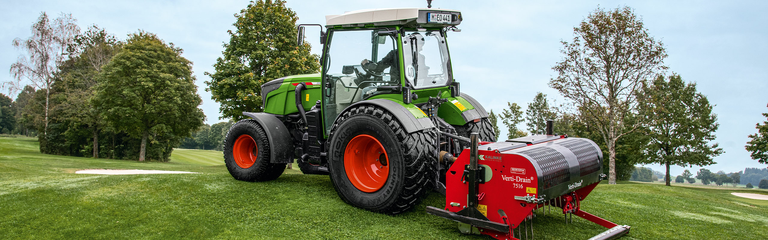 Der grüne Fendt 200 V/F/P Vario mit den roten Felgen steht mit einem roten Anbaugerät auf einem Hügel. Im Hintergrund sind grüne Bäume zu erkennen.