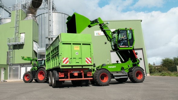 Fendt Cargo T955 mit Schaufel Ein Fendt Teleskoplader mit ausgefahrener Kabine und ausgeklappter Schaufel steht rechts neben einem grünen Anhänger vor einer grünen Lagerhalle und einem silbernen Silo.