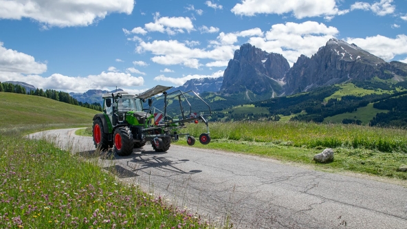 Ein grüner Fendt Tractor mit dem grünen Anbaugerät Fendt Former 351 DS, in eingeklappten Zustand, fährt auf einer schmalen grauen Straße. Im Hintergrund ist eine grüne Wiese, eine Berglandschaft und ein blauer Himmel mit weißen Wolken.