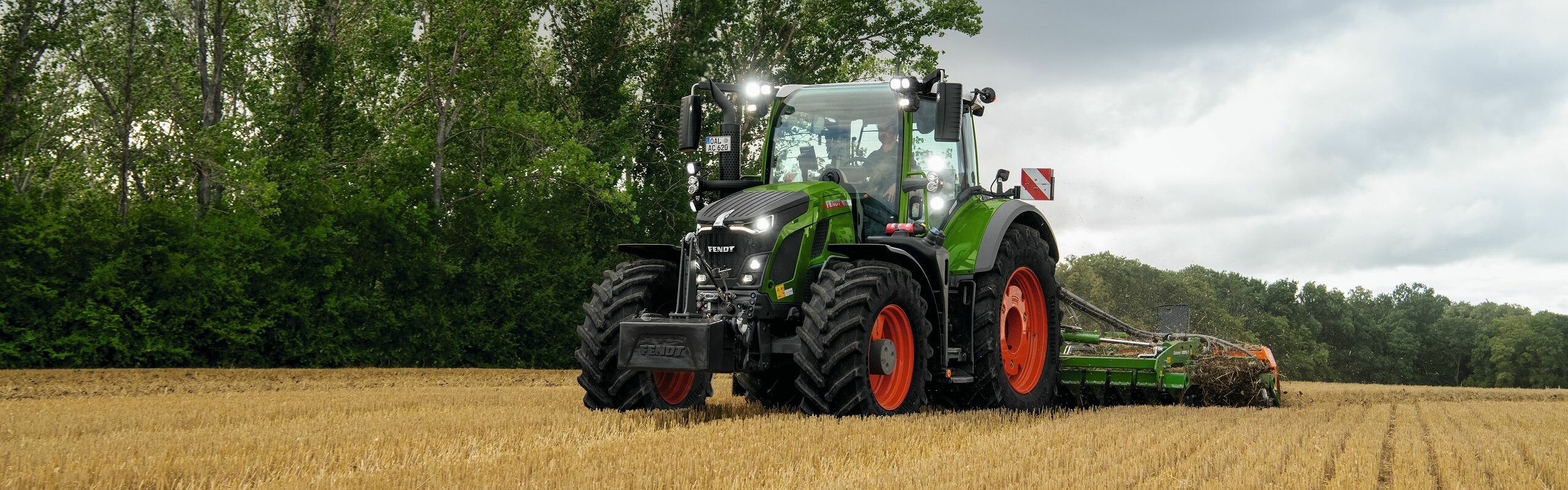 Der grüne Fendt 600 Vario Traktor mit roten Felgen im Einsatz auf einem Acker mit Bäumen im HIntergrund und einem wolkenbehangenen Himmel.