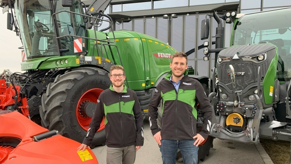 Zwei Fendt Mitarbeiter stehen vor einem Fendt Katana und einem Fendt Traktor. Im Hintergrund ist die Glasfassade eines Gebäudes zu erkennen.