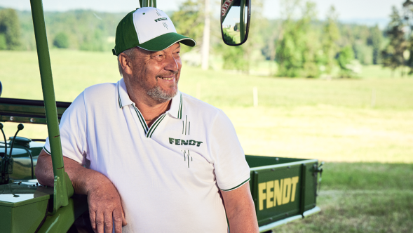 Ein lächelnder Mann steht vor einem grünen Fahrzeug mit Anhänger. Der Anhänger hat einen gelben Fendt-Schriftzug. Der Mann trägt ein weißes Polo-Shirt mit Fendt Logo und eine weiß-grüne Schirmmütze mit Fendt Logo aus dem Fendt Shop.