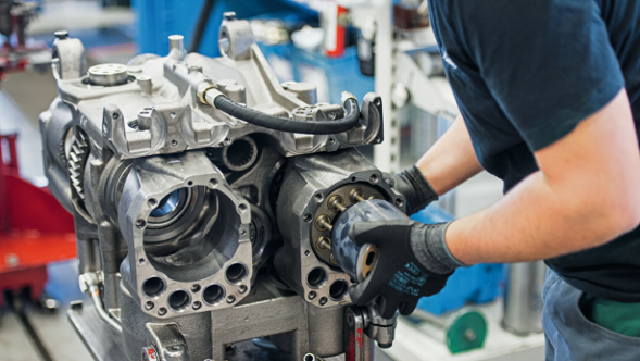 Ein Mechaniker arbeitet in einer Werkstatt an einem teilweise zerlegten Motorblock. Der Mechaniker trägt schwarze Handschuhe und hält ein rundes Bauteil, das er in den Motorblock einsetzt.