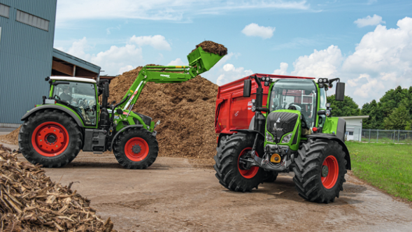 Das Bild zeigt zwei grüne Fendt Traktoren mit roten Felgen im Einsatz. Der linke Traktor hat einen Fendt Cargo Frontlader angebaut und lädt einen großen Haufen Mulch in einen roten Anhänger, der an den rechten Traktor angehängt ist.