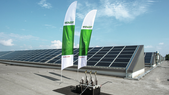 Auf dem Dach sind mehrere Reihen an Solarpanels, die in einem 45 Grad Winkel zum Boden stehen, installiert. Im Vordergrund stehen zwei weiß-grüne Flaggen mit dem Fendt-Logo. Im Hintergrund ist der blaue Himmel zu sehen.