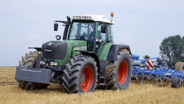 Ein grüner Fendt-Traktor mit weißem Dach zieht ein blaues Bodenbearbeitungsgerät über ein Feld. Im Hintergrund sind ein grüner Baum und der blaue Himmel zu sehen.