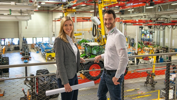Zwei Personen stehen in einer Fendt Produktionshalle, eine hält ein zusammengerolltes Dokument. Im Hintergrund wird ein Fendt Traktor montiert und es sind Maschinen und Montageeinrichtungen zu sehen.