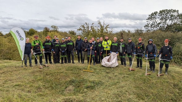 Eine Gruppe in Fendt Arbeitskleidung steht auf einer Wiese mit Werkzeugen wie Rechen und Sensen. Links neben der Gruppe steht eine BUND-Flagge. Im Hintergrund sind Bäume und Büsche zu sehen.