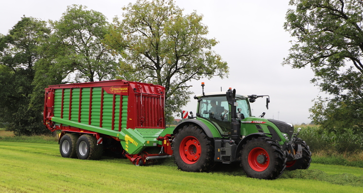 Ein Fendt Traktor fährt mit einem Strautmann Ladewagen im Gespann über eine Wiese und lädt Gras