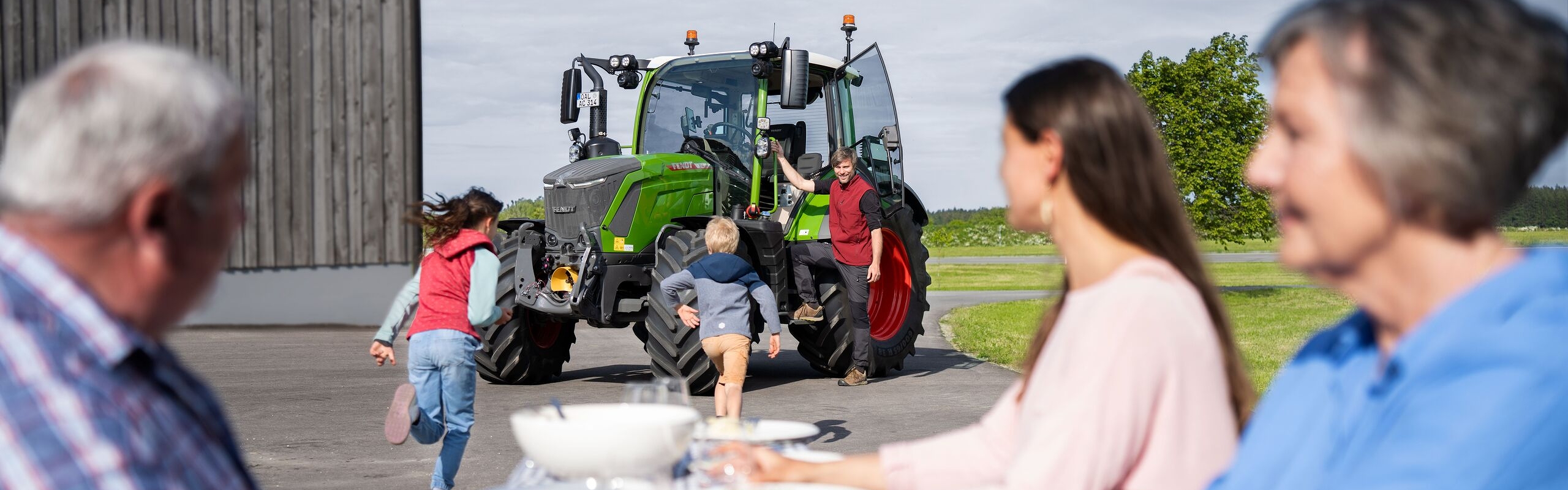 Ältere und jüngere Menschen sitzen gemeinsam am Tisch, während Kinder zu dem grünen Fendt Traktor 300 Vario Gen 5 laufen. Aus dem grünen Traktor steigt ein junger Mann aus.