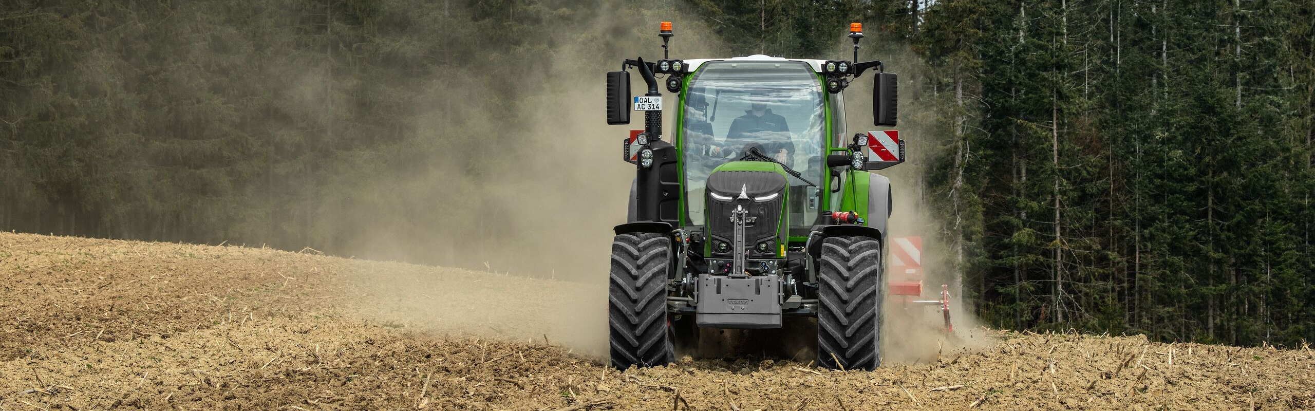 Ein Fendt 300 Vario Gen5 Traktor fährt über ein staubiges Feld vor einem Wald und wirbelt Erde auf.