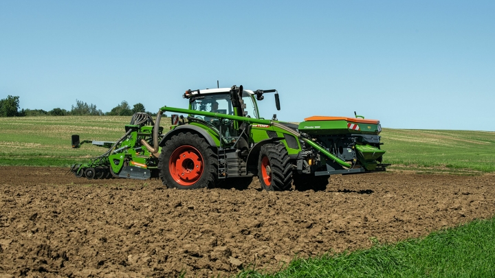 Der grüne Fendt 600 Vario mit roten Felgen und einer Drillmaschine als Anbaugerät fährt auf einem Acker. Im Hintergrund ist eine grüne Wiese und ein blauer Himmel zu sehen.