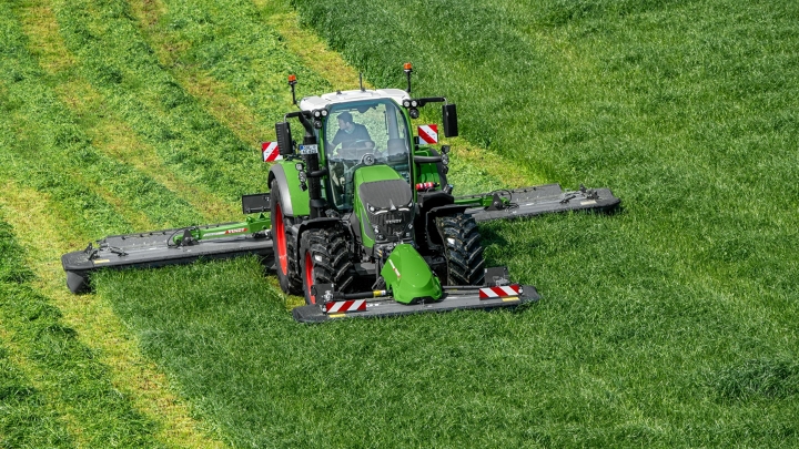 Der grüne Fendt 600 Vario mit roten Felgen und einer Drillmaschine als Anbaugerät fährt auf einem Acker. Im Hintergrund ist eine grüne Wiese und ein blauer Himmel zu sehen.