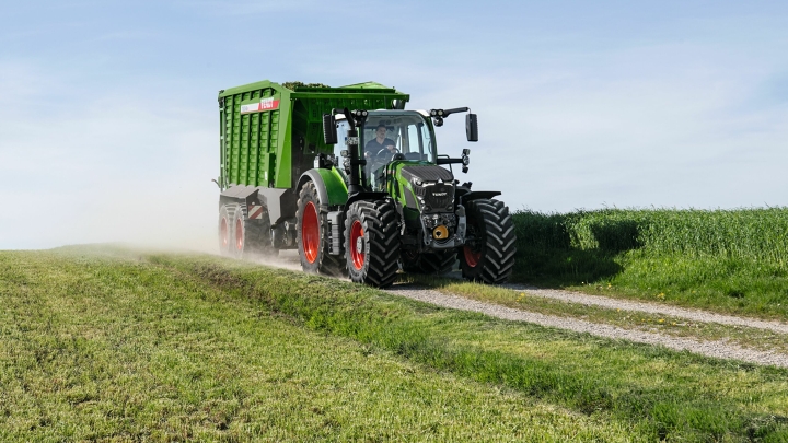 Der grüne Fendt 600 Vario Traktor mit roten Felgen und dem grünen Ladewagen Fendt Tigo fährt auf einer kleinen Feldstraße mit einer grünen Wiese im Vorder- und Hintergrund.