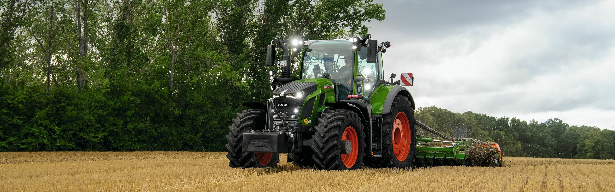 Der grüne Fendt 600 Vario Traktor mit roten Felgen im Einsatz auf einem Acker mit Bäumen im HIntergrund und einem wolkenbehangenen Himmel.